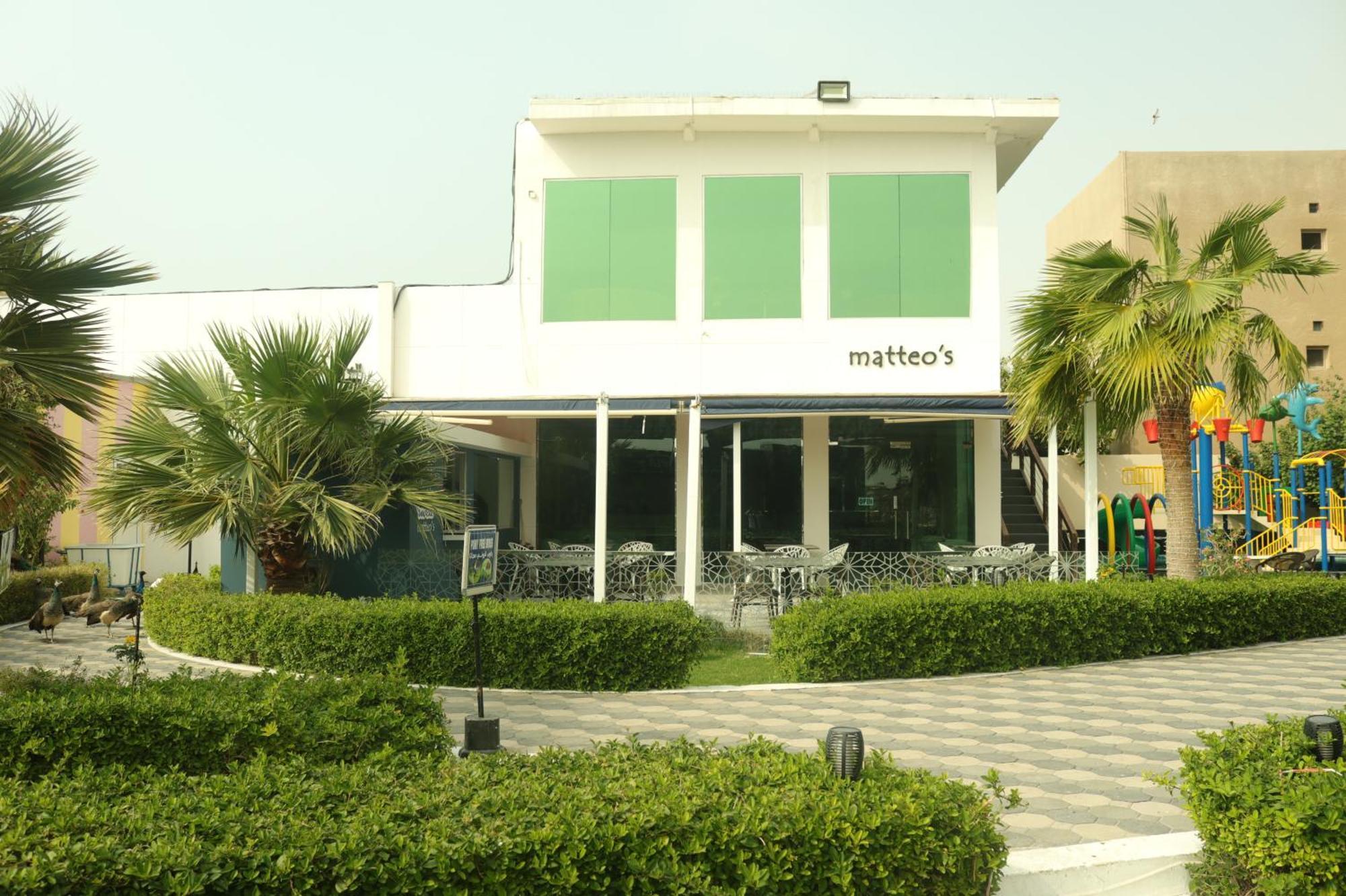 Lagoona Beach Luxury Resort And Spa Manama Eksteriør bilde The photo shows a building with the name "matteo's" displayed prominently on the front. The structure features large windows and green shutters. There is a landscaped area in front with bushes and palm trees, along with a paved pathway leading up to 