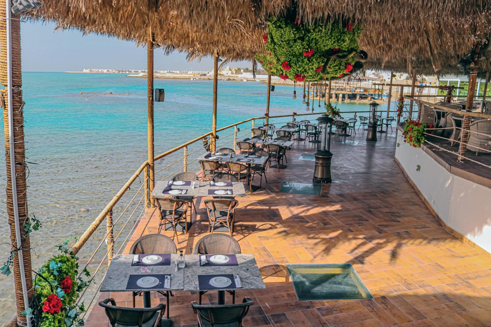 Lagoona Beach Luxury Resort And Spa Manama Eksteriør bilde The photo shows a seaside restaurant with a spacious outdoor dining area. It features several tables set with plates and utensils, surrounded by lush greenery and decorative plants. The flooring is made of brown tiles, and there are thatched roofs pr