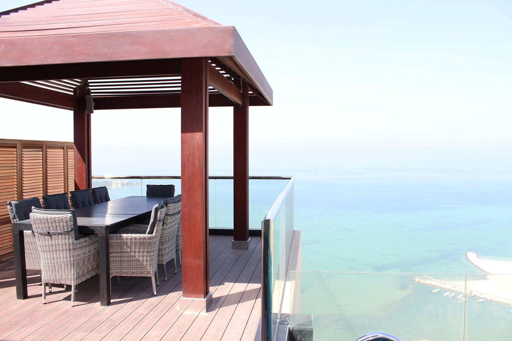 Lagoona Beach Luxury Resort And Spa Manama Eksteriør bilde The photo showcases a stylish outdoor patio or terrace area. It features a wooden structure with a sloped roof, providing a shaded space. There is a large table made of dark wood, surrounded by several wicker chairs. The patio is elevated and has gla