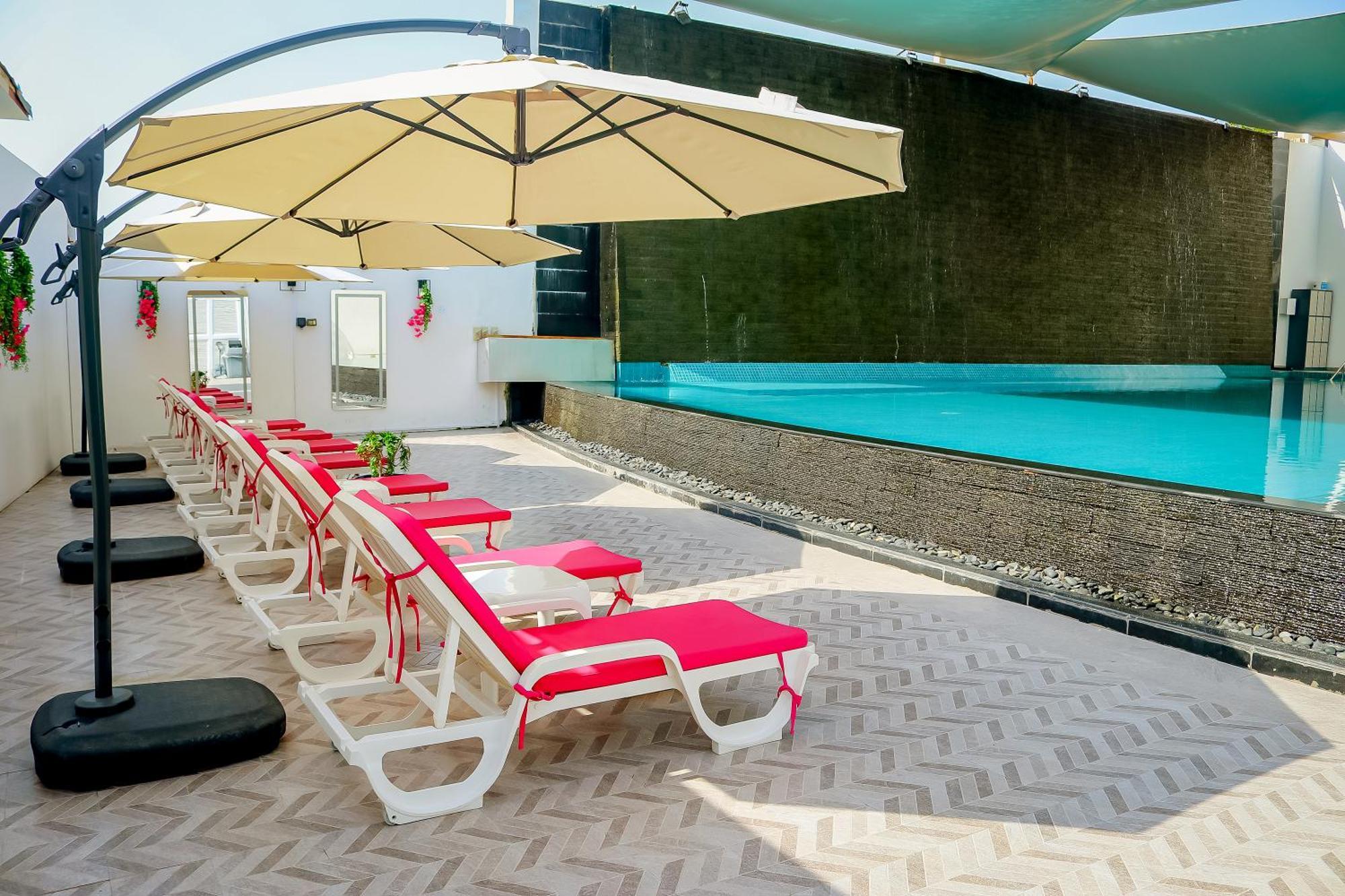 Lagoona Beach Luxury Resort And Spa Manama Eksteriør bilde The photo shows a sunny poolside area with several lounge chairs arranged in a row. Each chair features red cushions and is positioned under large, beige umbrellas for shade. In the background, there is a beautiful swimming pool with clear blue water