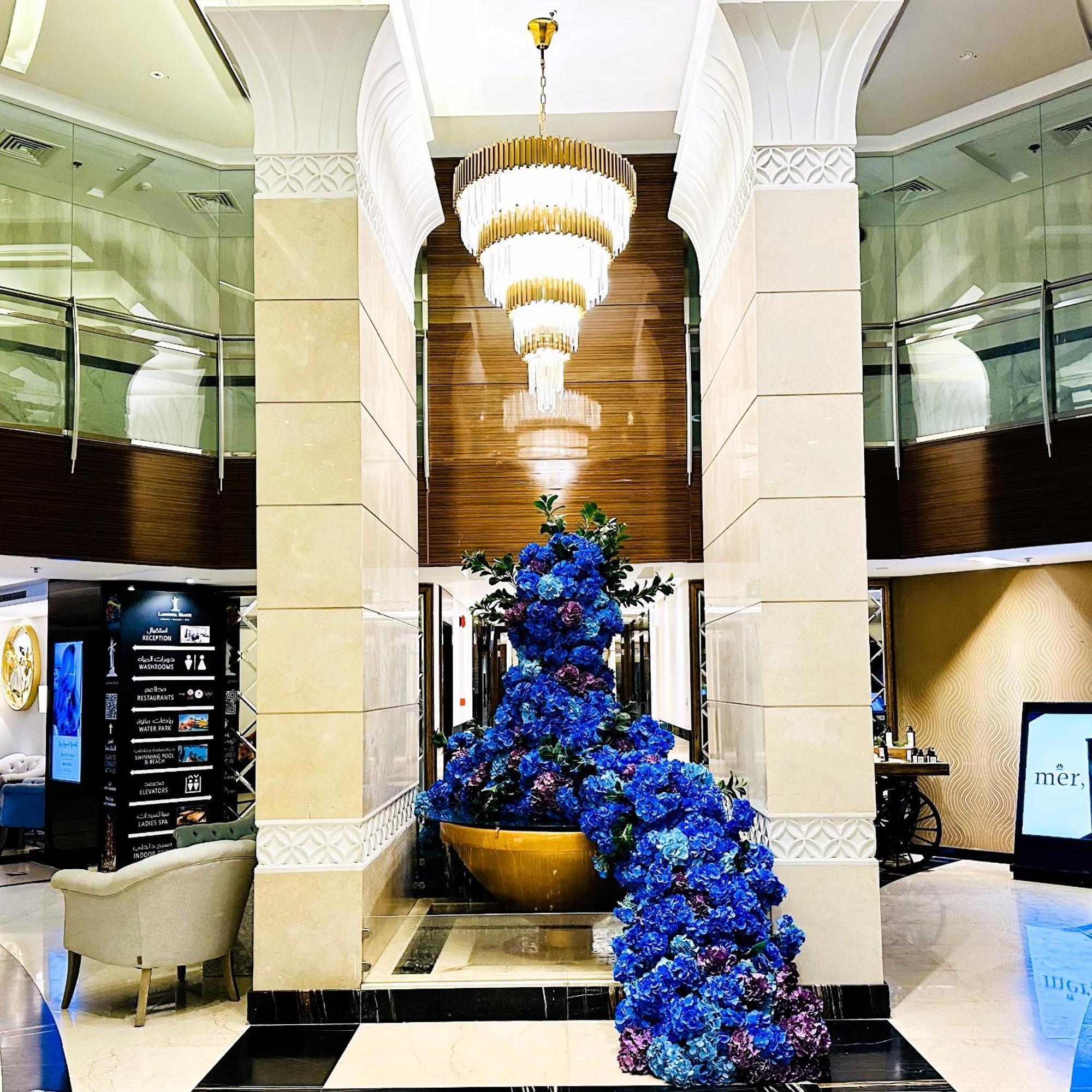 Lagoona Beach Luxury Resort And Spa Manama Eksteriør bilde The photo depicts an elegant lobby or entrance area of a building. Central to the scene is a large floral arrangement featuring vibrant blue flowers. This arrangement is positioned in a decorative bowl. Above it hangs a stylish chandelier with a laye