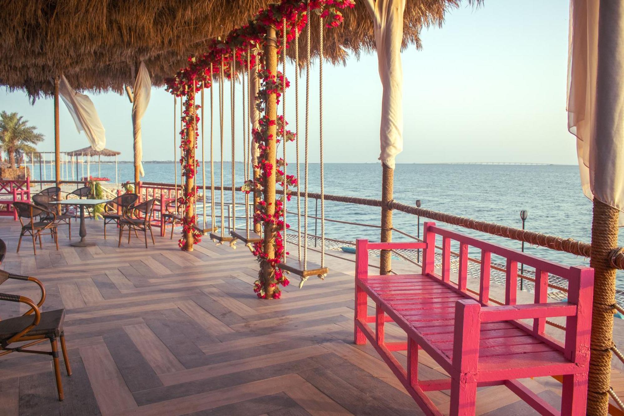 Lagoona Beach Luxury Resort And Spa Manama Eksteriør bilde The photo depicts a scenic outdoor area by the water, with a wooden deck and a thatched roof. Two swings adorned with flowers are hanging from the ceiling, providing a charming touch to the space. In the foreground, there is a bright pink bench, addi
