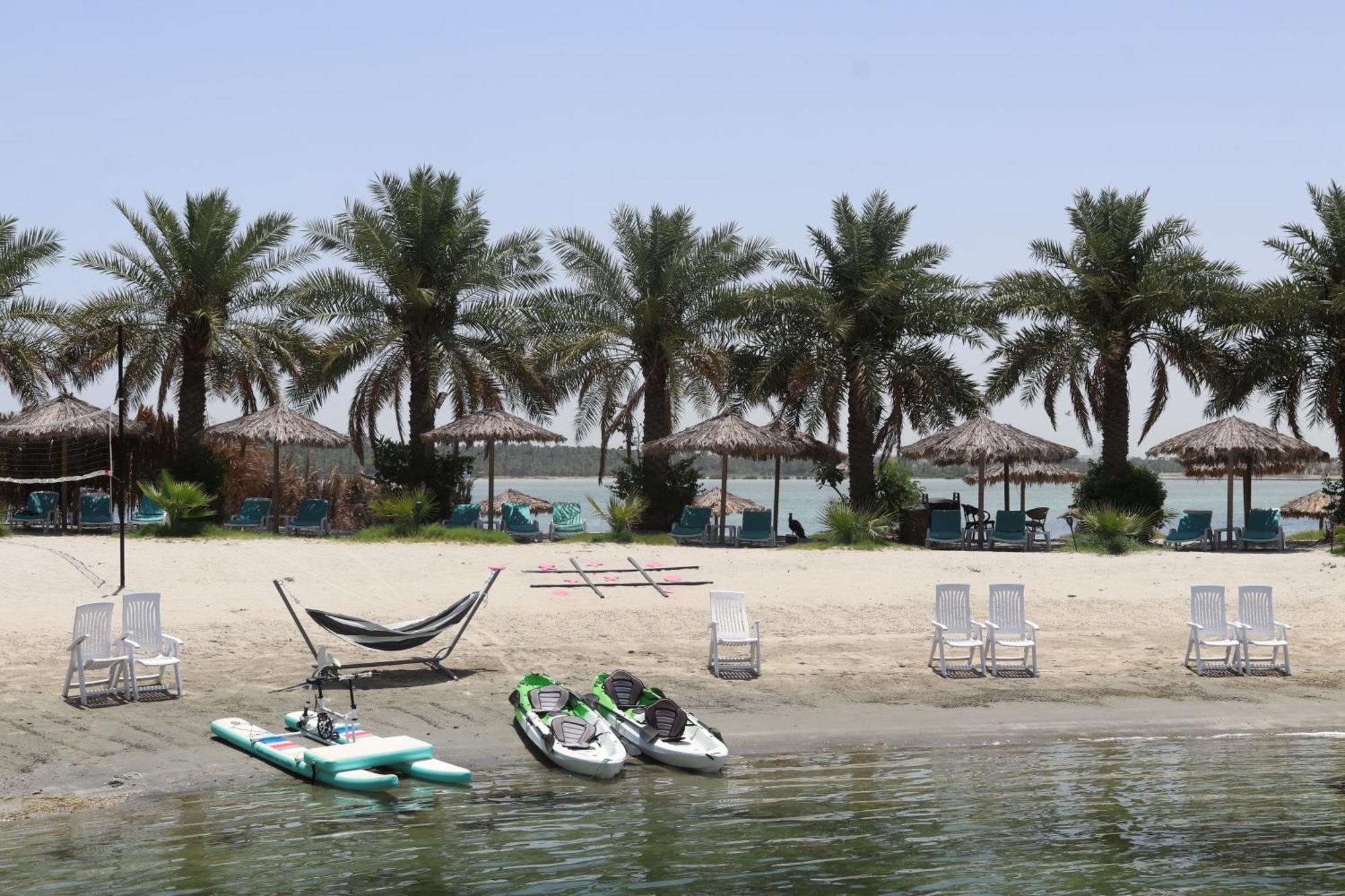 Lagoona Beach Luxury Resort And Spa Manama Eksteriør bilde The photo depicts a serene beach scene with several palm trees lining the shore. There are thatched umbrellas providing shade, and a few lounge chairs are positioned on the sand. A hammock is set up between two supports, inviting relaxation. In the f