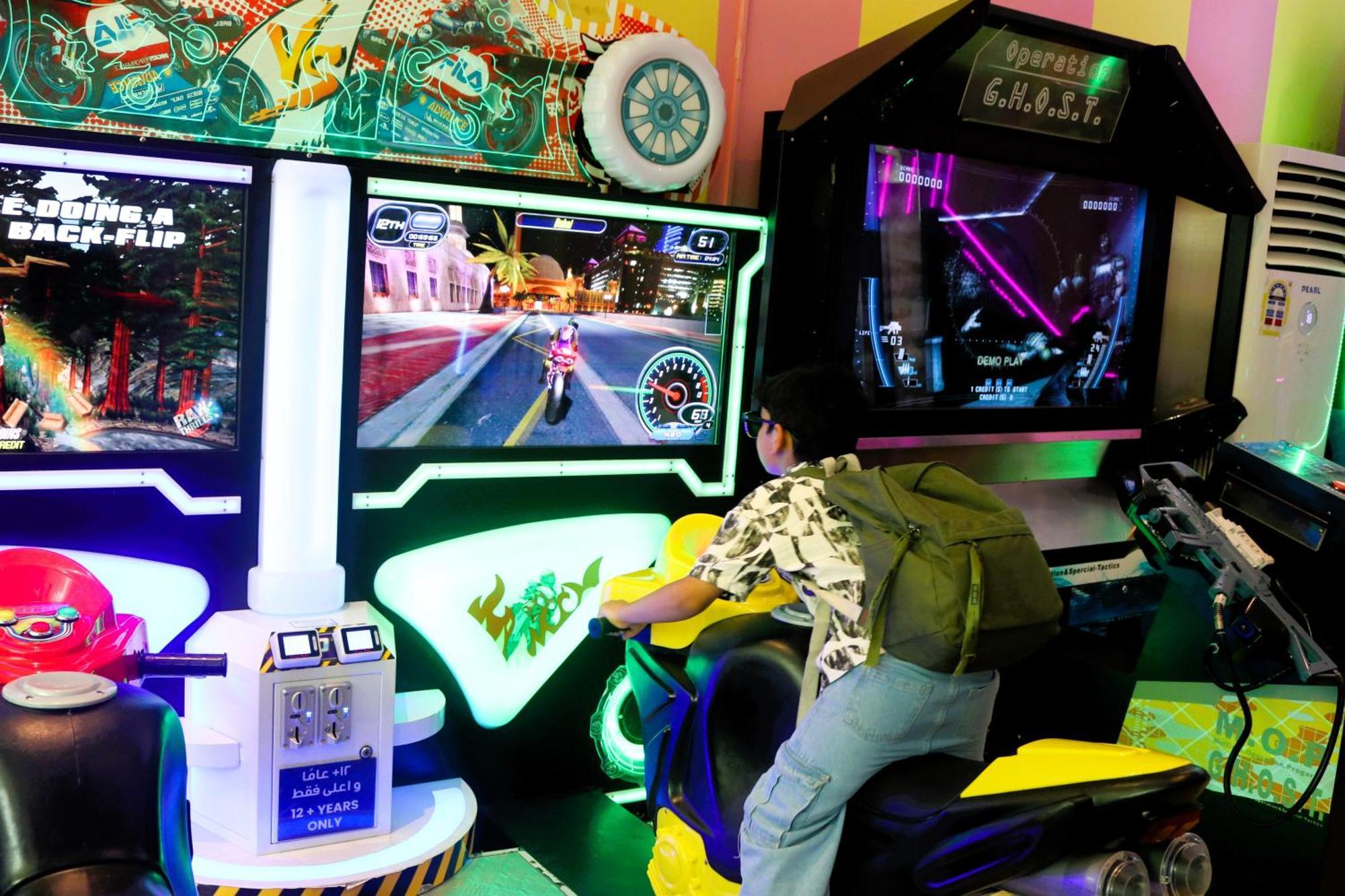 Lagoona Beach Luxury Resort And Spa Manama Eksteriør bilde The photo shows a person playing an arcade motorcycle racing game. They are seated on a motorcycle-shaped seat, actively engaging with the game displayed on the screen in front of them. The screen shows a vibrant racing scene with streets and colorfu