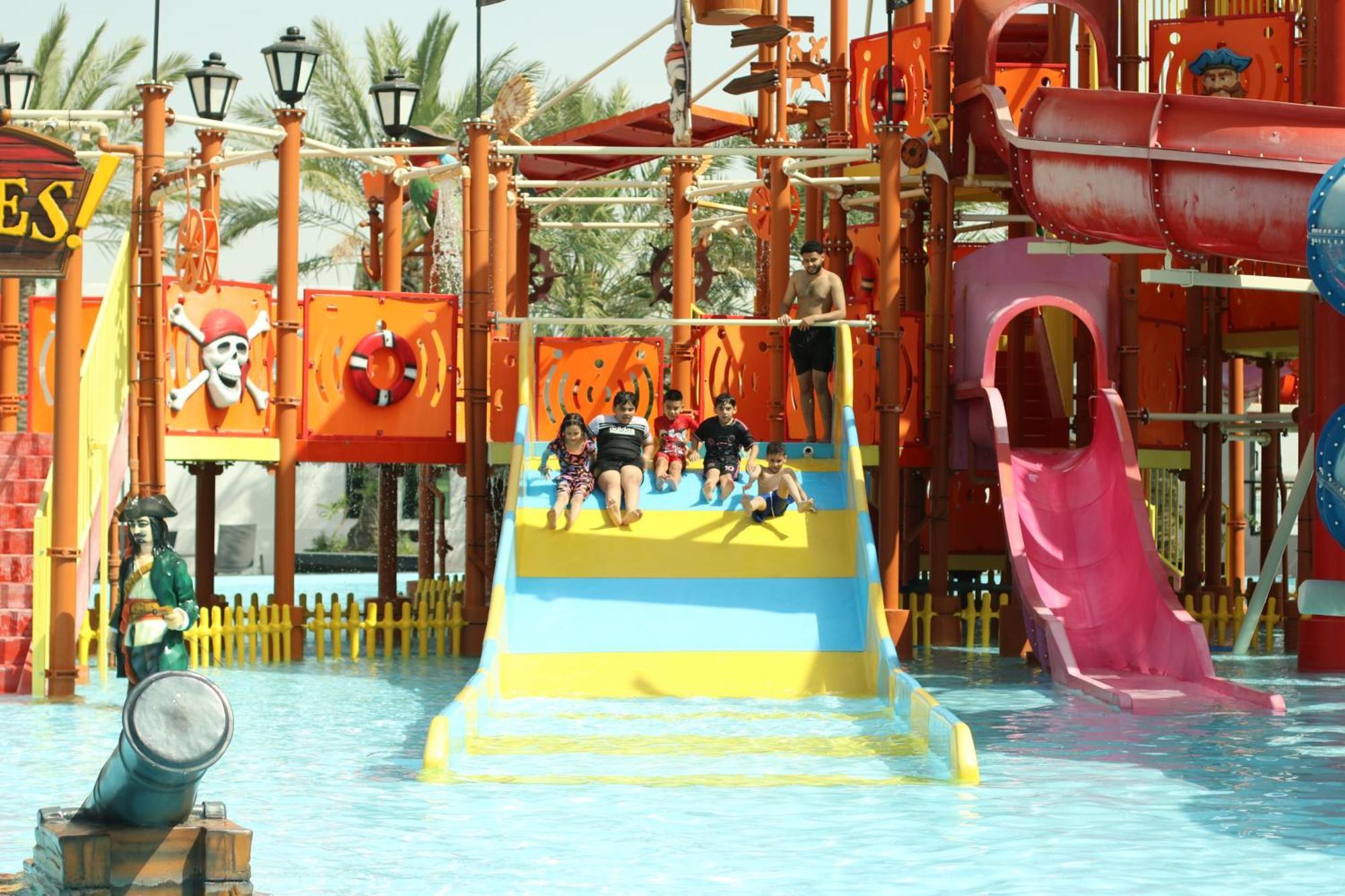 Lagoona Beach Luxury Resort And Spa Manama Eksteriør bilde The photo shows a colorful water park scene featuring a play area with multiple slides. There are children enjoying themselves, sliding down bright yellow and pink slides into a pool of water. The structure has pirate-themed decorations, including a 