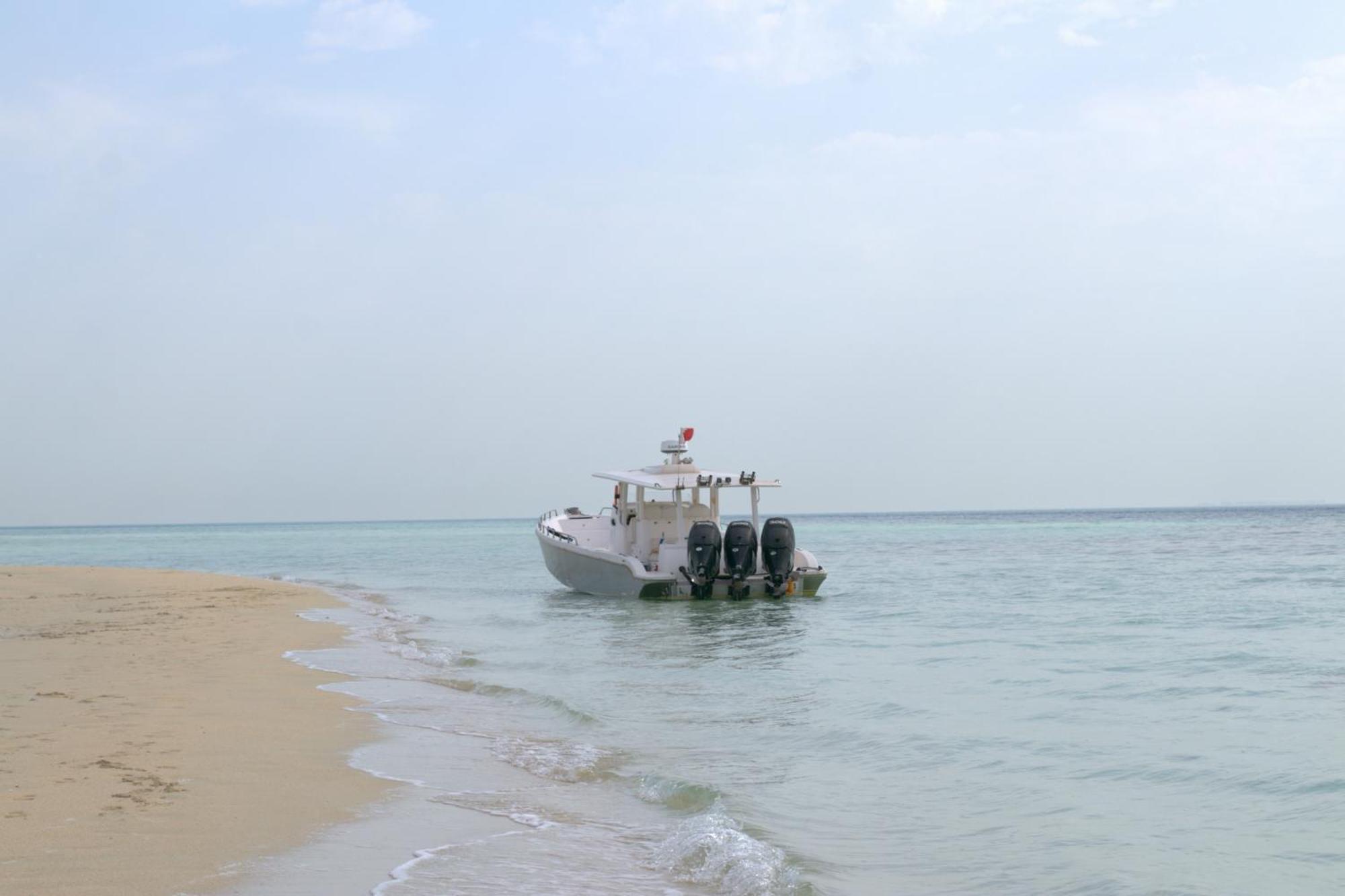 Lagoona Beach Luxury Resort And Spa Manama Eksteriør bilde The photo depicts a small boat anchored near a sandy beach, partially submerged in calm, clear water. The boat is white and features three outboard motors on its stern. In the background, the sea stretches out toward the horizon under a light, cloudy