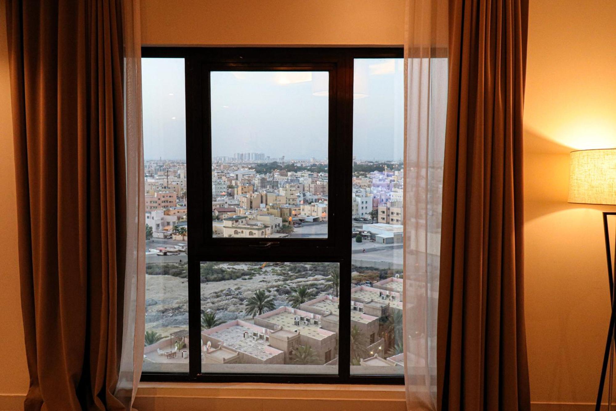 Lagoona Beach Luxury Resort And Spa Manama Eksteriør bilde The photo shows a window with a black frame and sheer curtains. Through the window, there is a view of a cityscape, displaying a densely populated area with various buildings and rooftops. The sky appears slightly overcast, suggesting a muted lightin