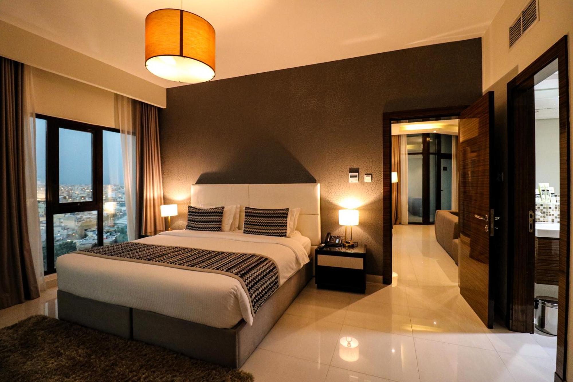 Lagoona Beach Luxury Resort And Spa Manama Eksteriør bilde The photo depicts a modern hotel room or suite. It features a large bed with a white and gray color scheme, accented with decorative pillows. On either side of the bed, there are sleek bedside lamps providing warm lighting. The walls are a dark color