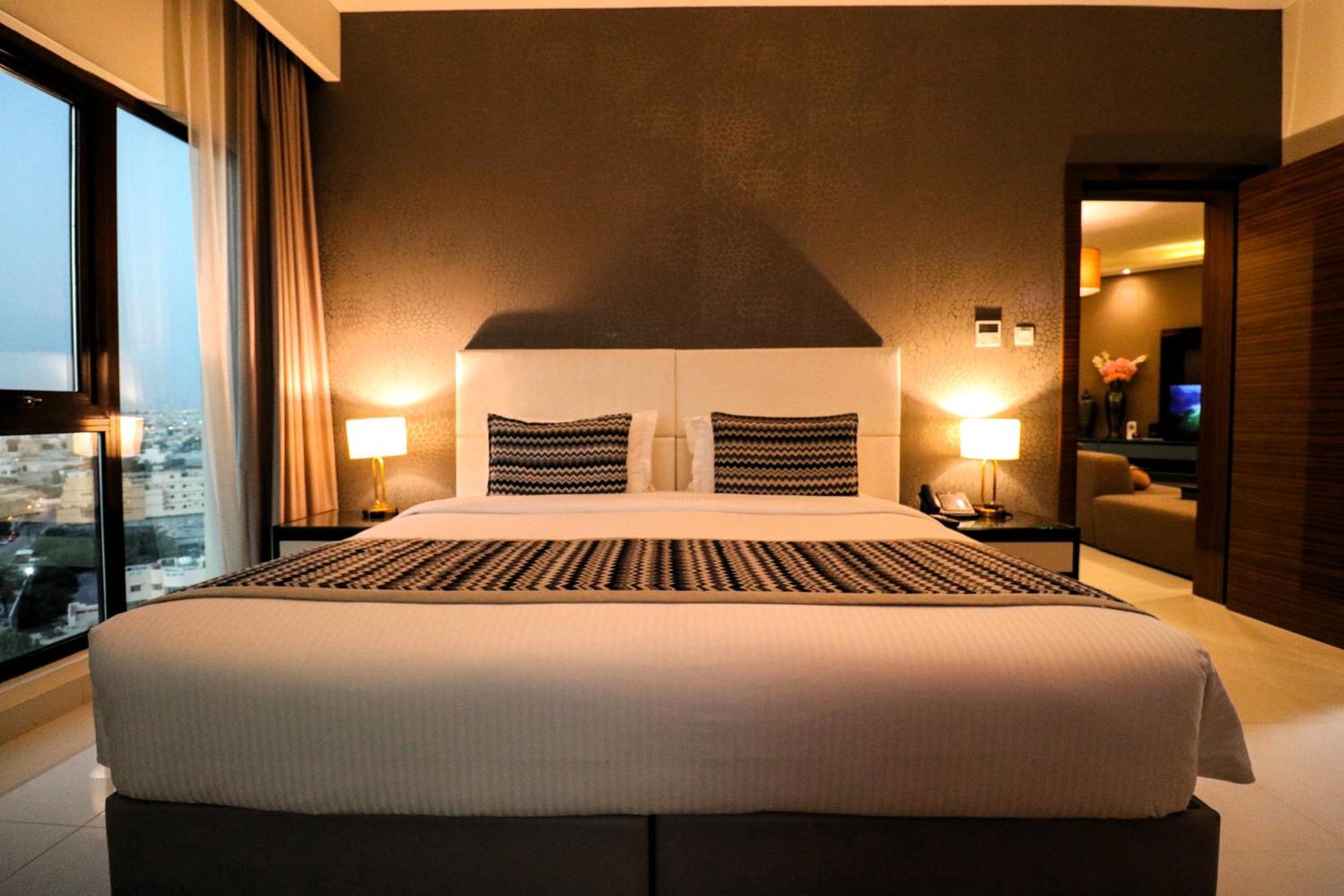 Lagoona Beach Luxury Resort And Spa Manama Eksteriør bilde The photo depicts a modern hotel room featuring a large bed with a white comforter and two decorative pillows. The headboard is light-colored, and the walls are painted a dark hue. On either side of the bed, there are bedside tables with lamps. The r