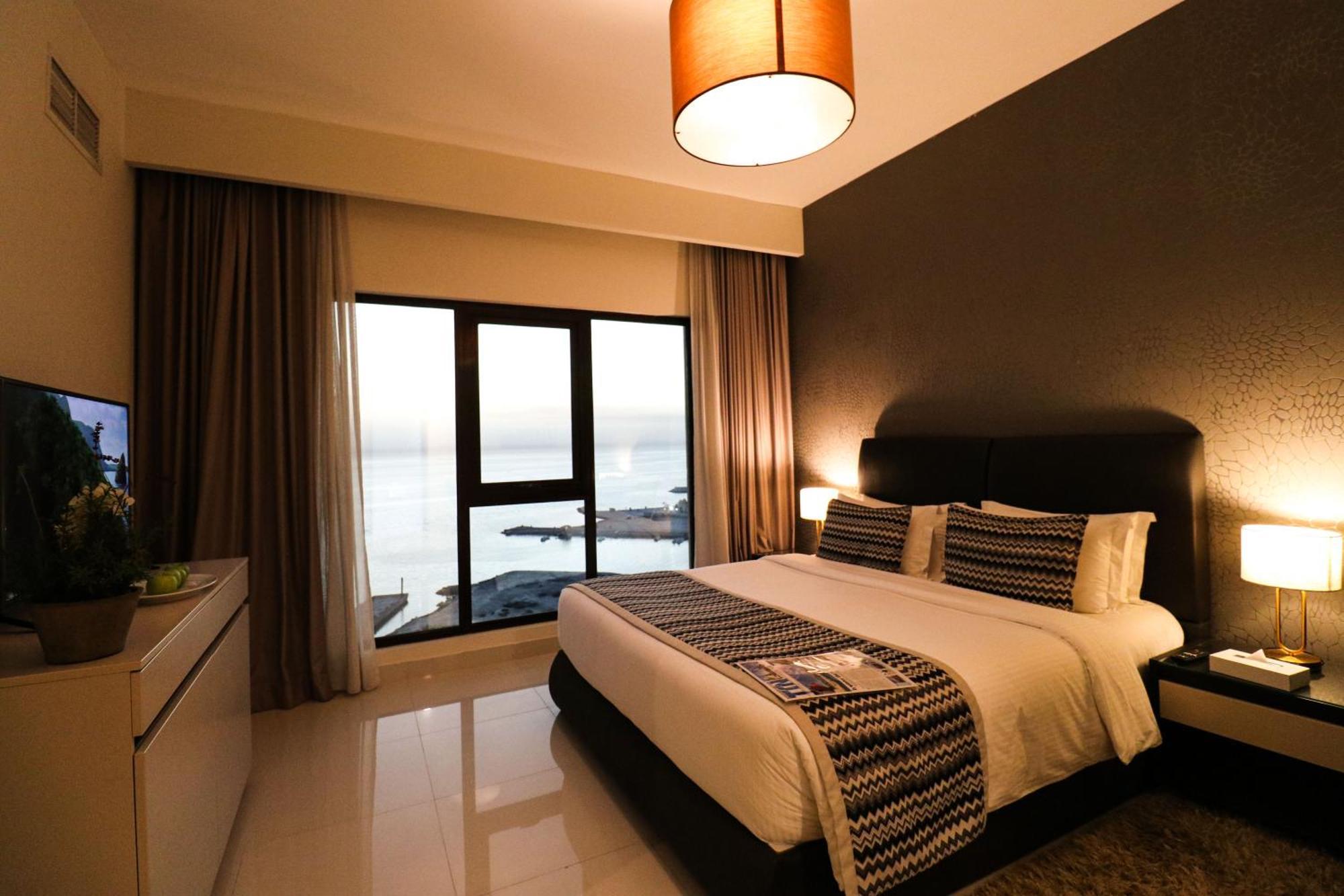 Lagoona Beach Luxury Resort And Spa Manama Eksteriør bilde The photo shows a modern hotel room featuring a neatly made bed with decorative pillows. The room has a stylish design with a dark accent wall and a large window that offers a view of the outdoors. Natural light comes in, giving the space a bright an