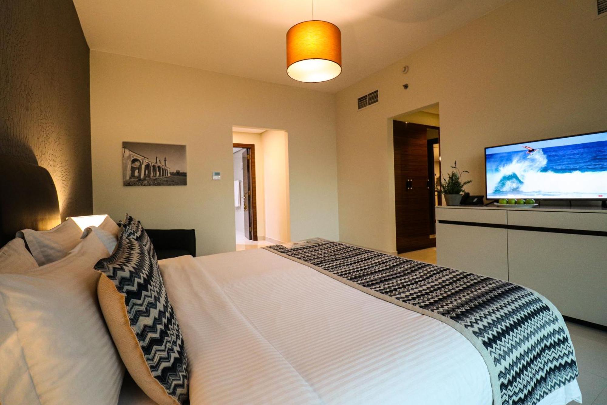 Lagoona Beach Luxury Resort And Spa Manama Eksteriør bilde The photo shows a well-decorated hotel room. There is a large bed with white linens and decorative pillows featuring a zigzag pattern. On the wall beside the bed is a piece of artwork, and there is a modern television on a white furniture unit opposi