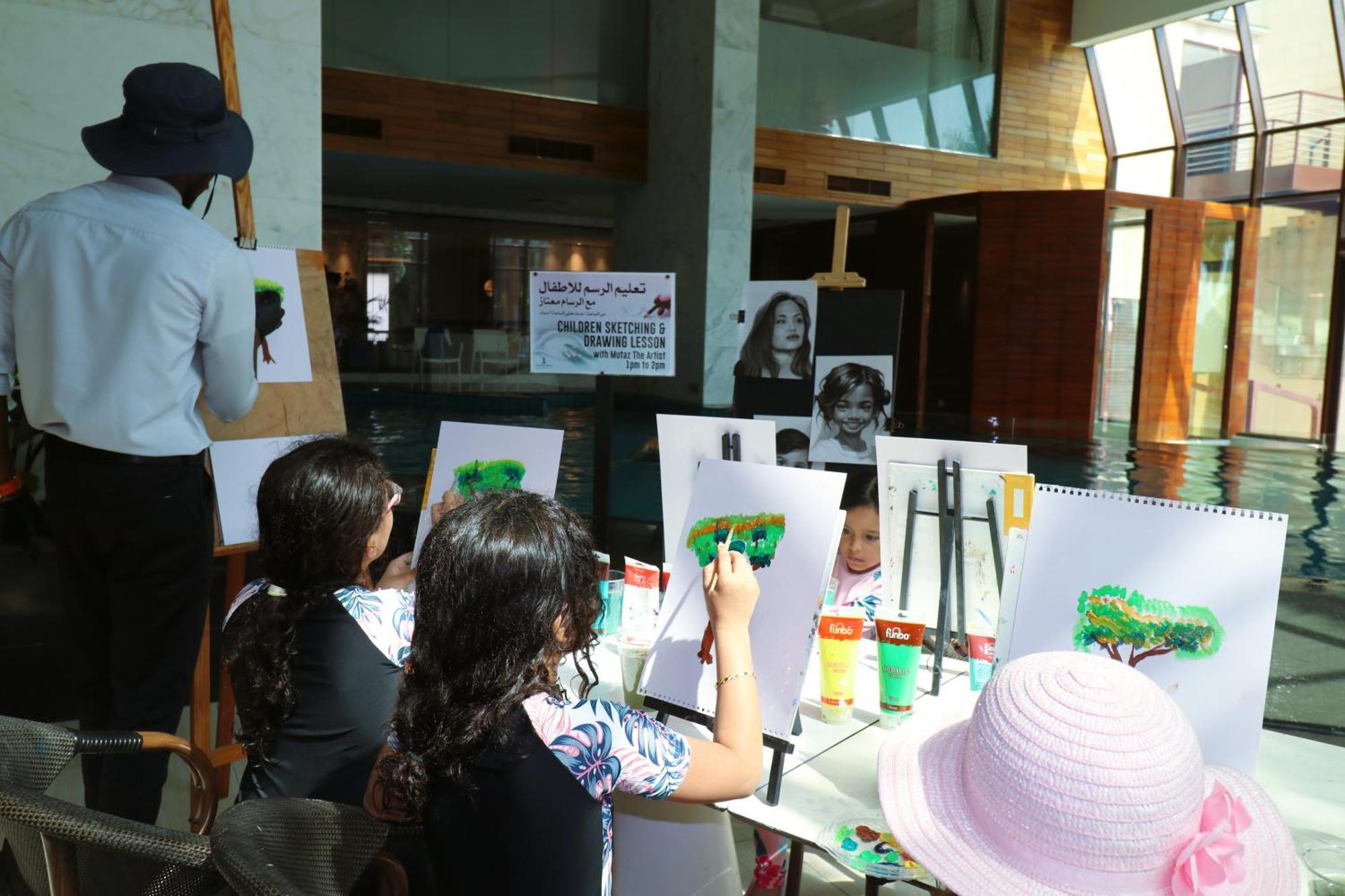 Lagoona Beach Luxury Resort And Spa Manama Eksteriør bilde The photo shows a group of children and an adult engaged in an art activity near a pool area. The children are seated at a table, painting on canvases or paper, with a focus on trees, as indicated by the green paint they are using. An adult, wearing 