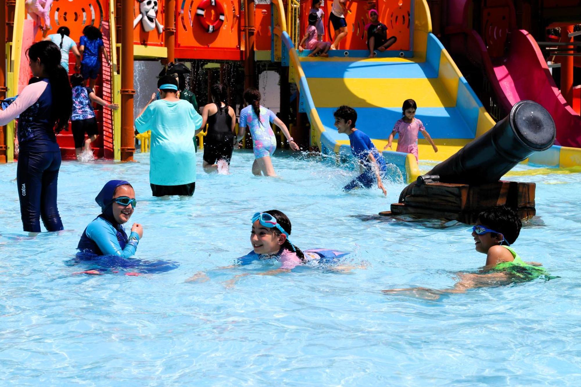 Lagoona Beach Luxury Resort And Spa Manama Eksteriør bilde The photo shows a vibrant water park scene with several children playing in a shallow pool. Some are swimming and splashing around, while others are playing on colorful slides in the background. The atmosphere is lively and cheerful, with kids of var