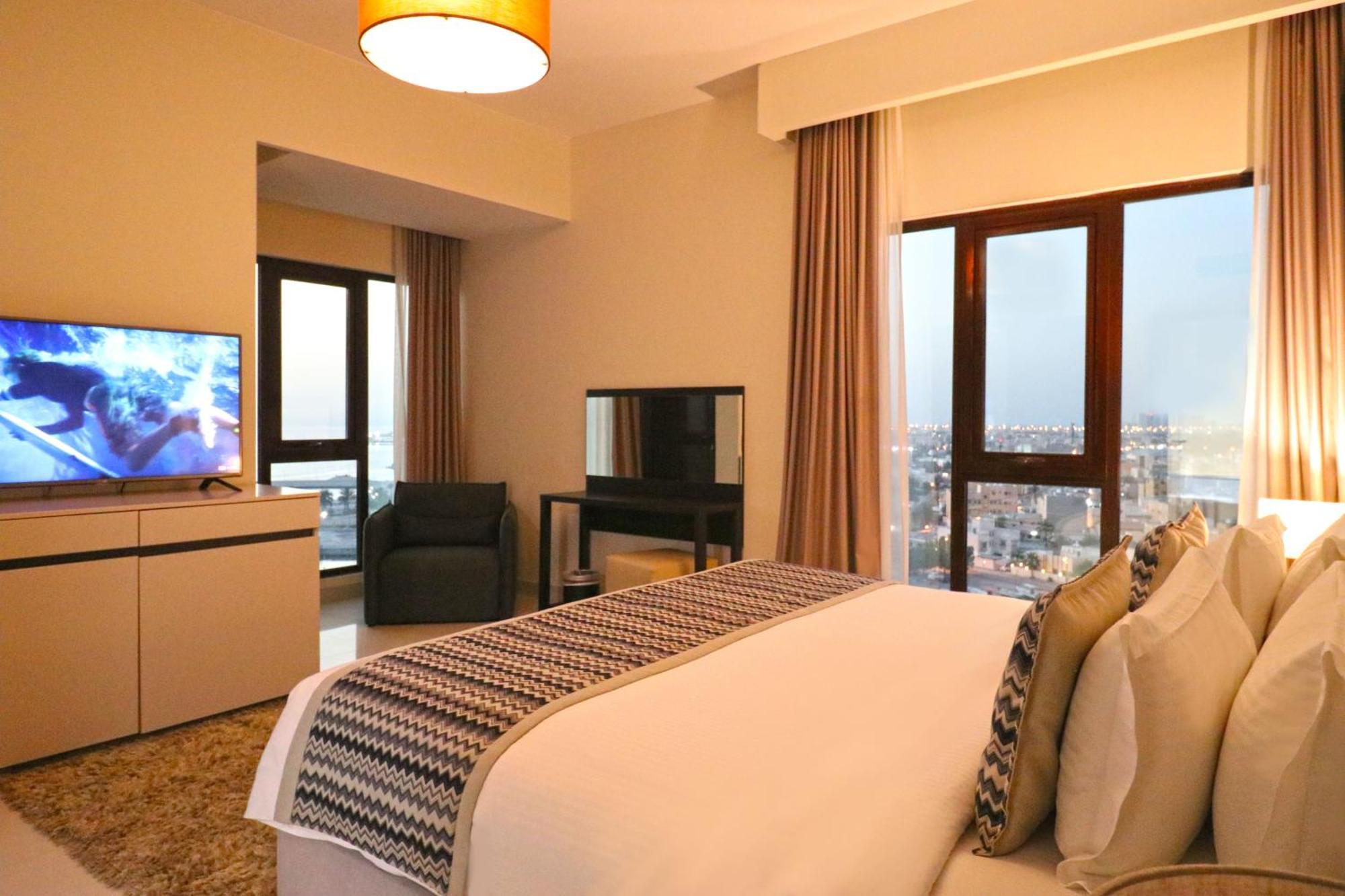 Lagoona Beach Luxury Resort And Spa Manama Eksteriør bilde The photo shows a modern hotel room or suite. It features a neatly made bed with decorative pillows in the foreground. To the left, there is a TV on a cabinet, and further back, a comfortable chair is situated in front of a large window that likely o