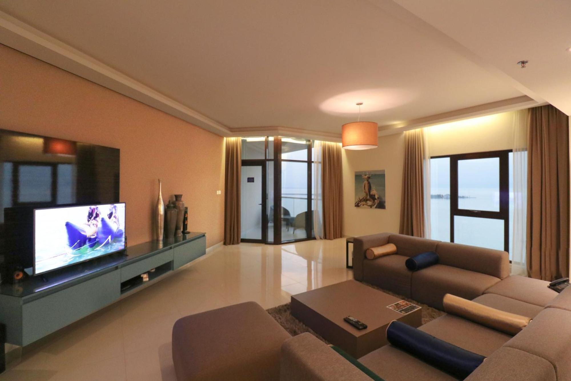 Lagoona Beach Luxury Resort And Spa Manama Eksteriør bilde The photo shows a modern living room with a sleek design. There is a large wall-mounted television on a contemporary entertainment unit, with decorative items placed beside it. The room features a comfortable sectional sofa in light tones, with a cof