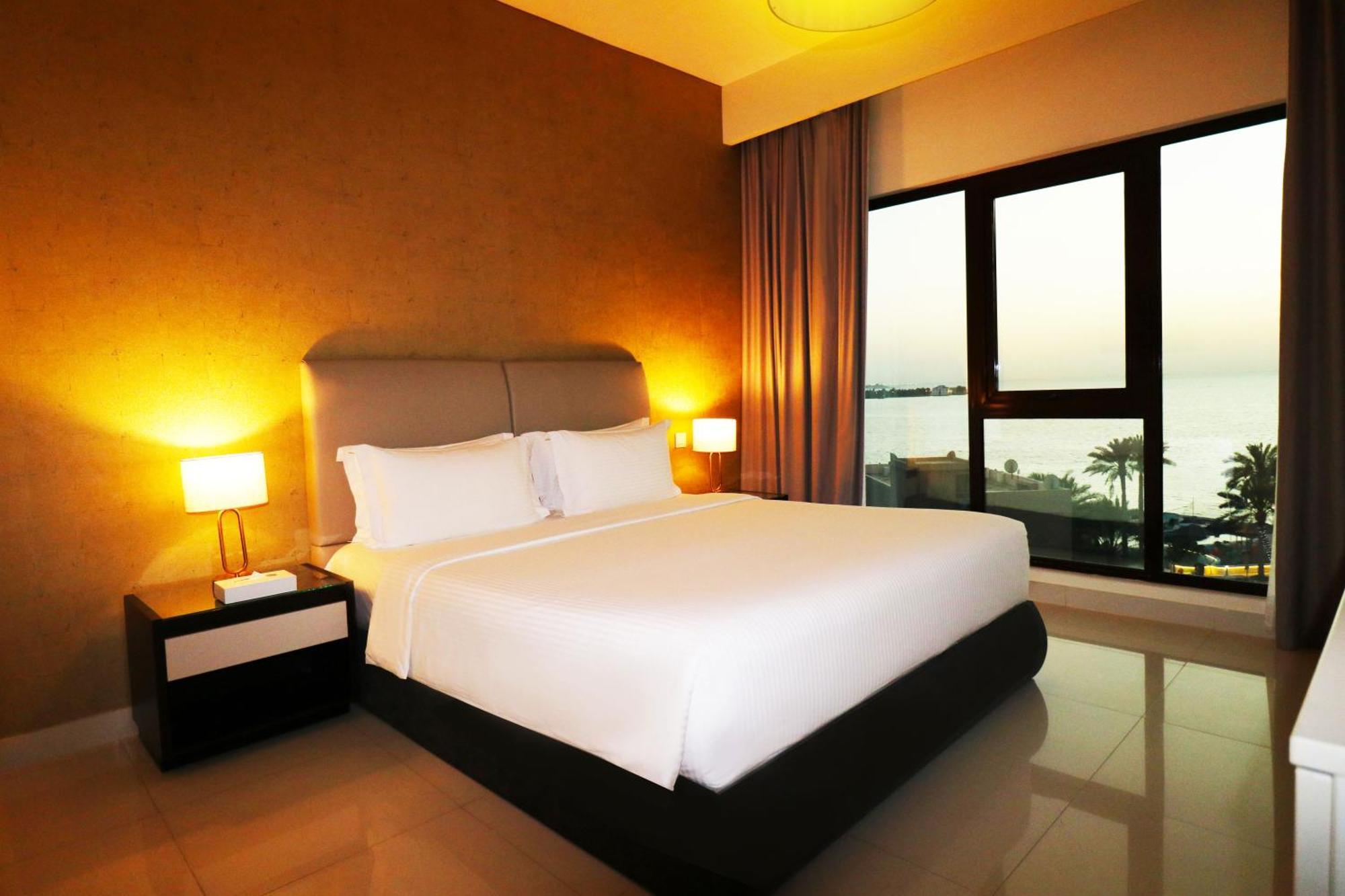Lagoona Beach Luxury Resort And Spa Manama Eksteriør bilde The photo shows a modern hotel bedroom with a large, comfortable bed dressed in white linens. The room features warm lighting, including bedside lamps, creating a cozy atmosphere. There are large windows with a view of the outdoors, possibly overlook