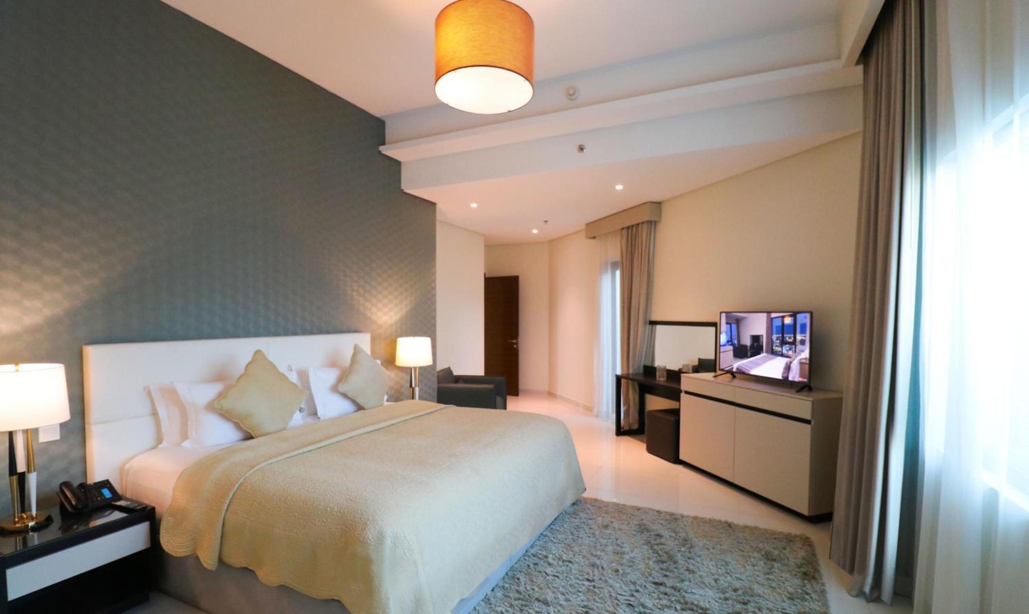 Lagoona Beach Luxury Resort And Spa Manama Eksteriør bilde The photo shows a modern hotel room featuring a large bed with a light-colored blanket and decorative pillows. The walls are painted in a subtle shade, and there is a textured accent wall behind the bed. On one side, a bedside lamp provides warm ligh