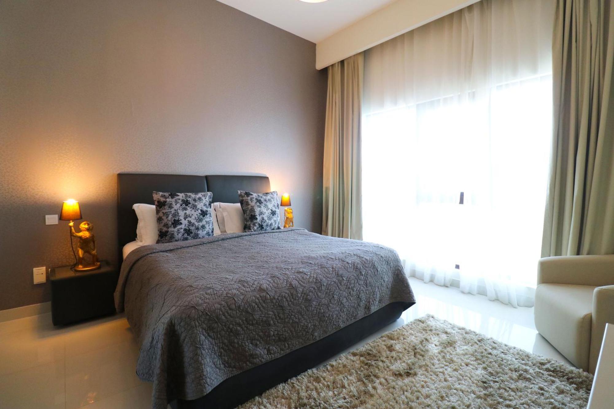 Lagoona Beach Luxury Resort And Spa Manama Eksteriør bilde The photo shows a stylish bedroom with a modern design. It features a large bed with a dark gray bedspread and decorative pillows. On either side of the bed are two wooden nightstands, each with a lamp. There is a large window covered with light, flo